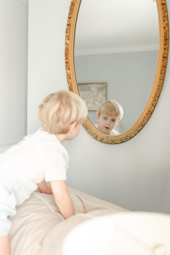 A blonde little boy peeks into a gold, grandmillenial style mirror on the way of his brother's nursery.