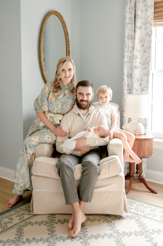 A family sits in a nursery rocking chair and smiles as dad holds the baby. A gold oval mirror is behind them.