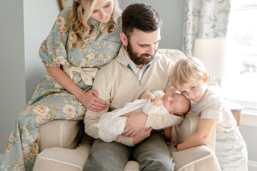 A toddler boy hugs his newborn brother as his dad holds him in a grandmillenial nursery rocking chair