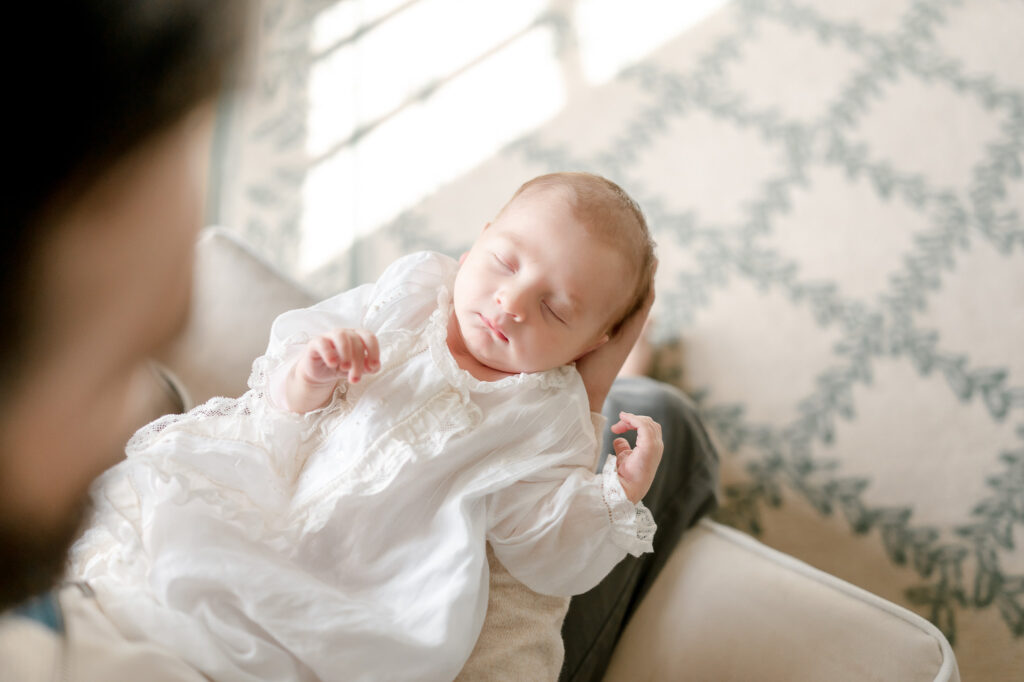 A newborn sleeps in his father's arms wearing a vintage infant gown.