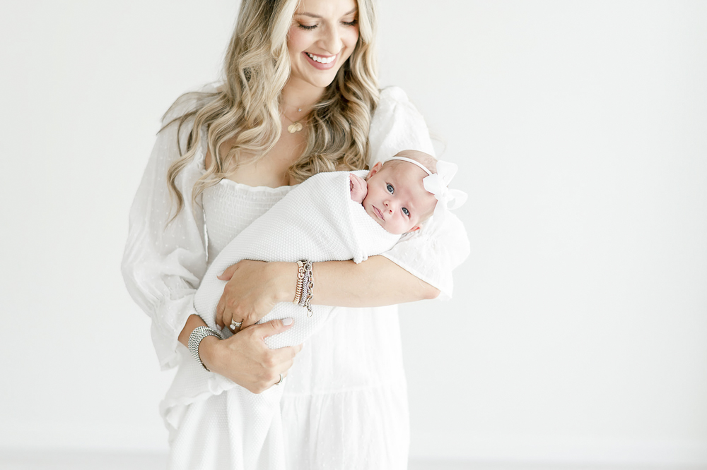 A mother wearing a white dresses holds her newborn in a white swaddle and white bow