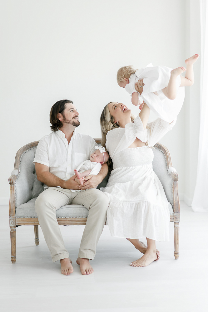 A new family of four sits on a bench and mom holds the toddler in the air as they all smile