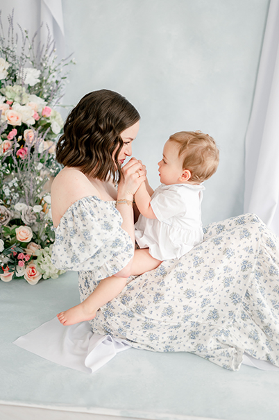 A mother in a blue floral dresses kisses her toddler son's hands