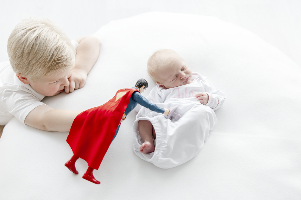 A little boy flies his superman doll over his newborn sister